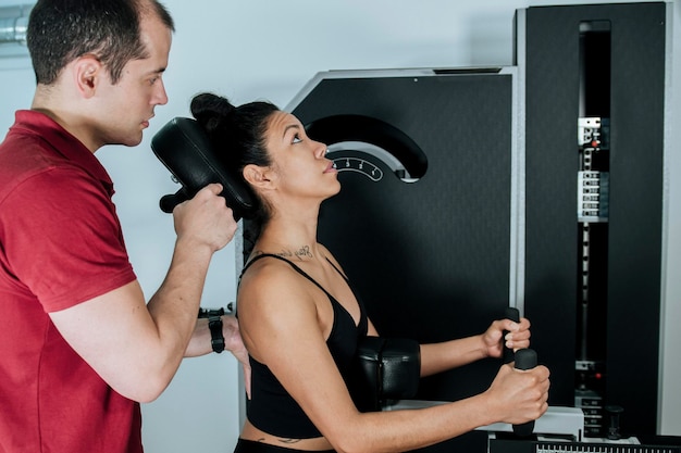 Junges athletisches Mädchen, das mit ihrem Personal Trainer ein Fitnessgerät benutzt, um den Halsbereich zu stärken