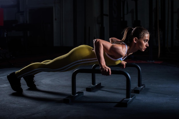 Junges athletisches Mädchen in gelber enger Sportuniform macht horizontale Pushups mit Stangen im Fitnessstudio