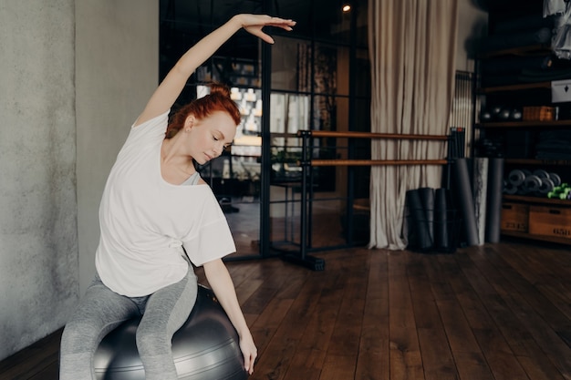 Junges athletisches rothaariges Mädchen in weißem übergroßem T-Shirt sitzt auf einem großen Fitball und macht Körperbeugen von einer Seite zur anderen vor dem Hintergrund der Fitnessgeräte. Pilates-Trainingskonzept