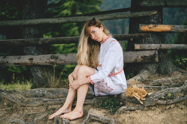 Foto junges attraktives barfuß blondes mädchen im weißen kleid mit verzierung, die nahe holzzaun draußen sitzt