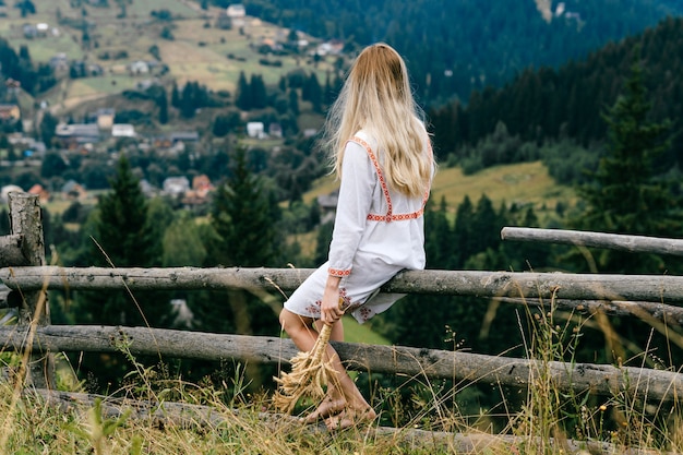 Junges attraktives blondes mädchen im weißen kleid mit verzierung, die auf holzzaun mit ährchenstrauß über malerischer landschaftslandschaft sitzt