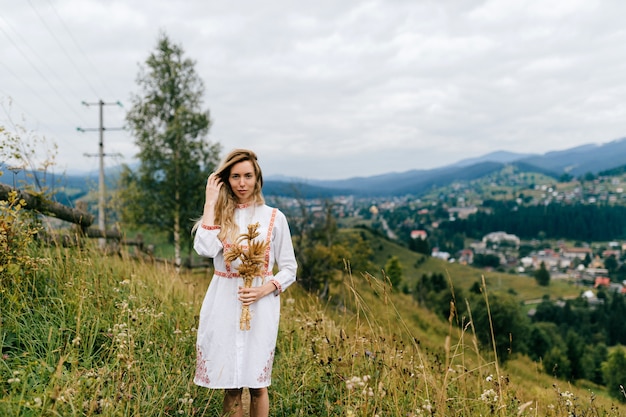 Junges attraktives blondes Mädchen im weißen Kleid mit Verzierung, die mit Ährchenstrauß über malerischer Landschaftslandschaft aufwirft