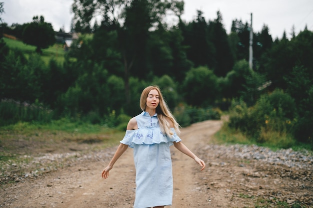 Junges attraktives elegantes blondes Mädchen im blauen Kleid, das auf der Straße in der Landschaft aufwirft