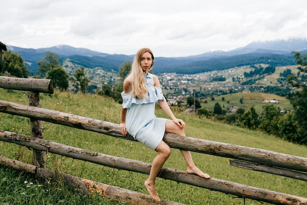 Foto junges attraktives elegantes blondes mädchen im blauen romantischen kleid, das auf dem hölzernen zaun in der landschaft sitzt
