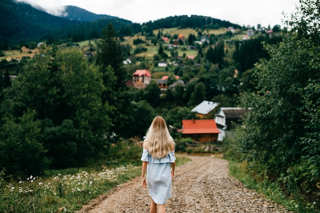 Junges attraktives elegantes blondes Mädchen im blauen romantischen Kleid, das zurück auf der Straße in der Landschaft aufwirft