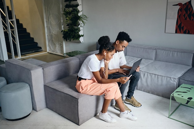 Junges attraktives lässiges afroamerikanisches Paar, das nachdenklich mit Laptop zusammen auf dem Sofa in einem modernen Co-Working-Space arbeitet