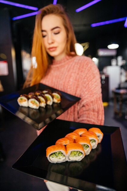 Junges attraktives Mädchen, das eine Makizushi-Sushi-Rolle hält, die auf schwarzem Teller auf Sushibar-Hintergrund serviert wird
