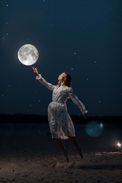 Junges attraktives Mädchen in einem langen weißen Kleid am Strand streckt ihre Arme zum Mond