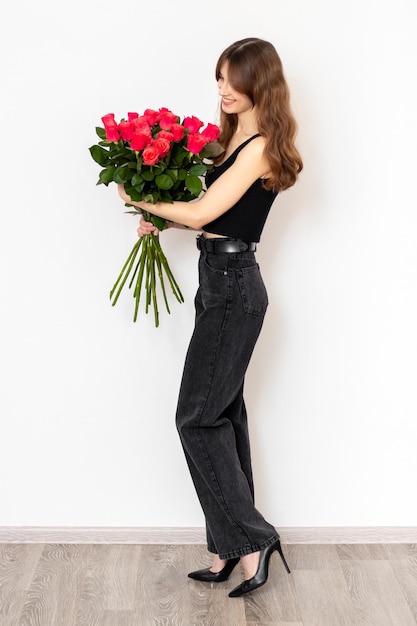 Junges attraktives Mädchen mit einem Strauß roter Rosen in vollem Wachstum auf weißem Hintergrund, Seitenansicht, das Konzept von Glück, Freude und Feier