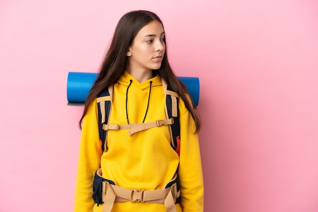 Junges Bergsteigermädchen mit einem großen Rucksack einzeln auf rosa Hintergrund, der zur Seite schaut