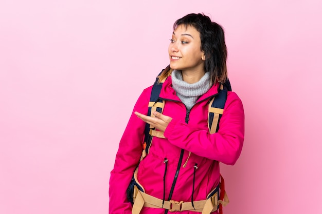 Junges Bergsteigermädchen mit einem großen Rucksack über isoliertem rosa Hintergrund, der Hände zur Seite für Einladung zum Kommen ausdehnt