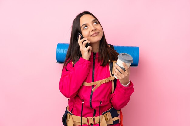 Junges Bergsteigermädchen mit einem großen Rucksack über isolierter rosa Wand, die Kaffee zum Mitnehmen und ein Handy hält