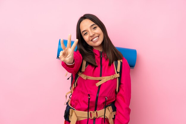 Junges Bergsteigermädchen mit einem großen Rucksack über isolierter rosa Wand glücklich und drei mit den Fingern zählend