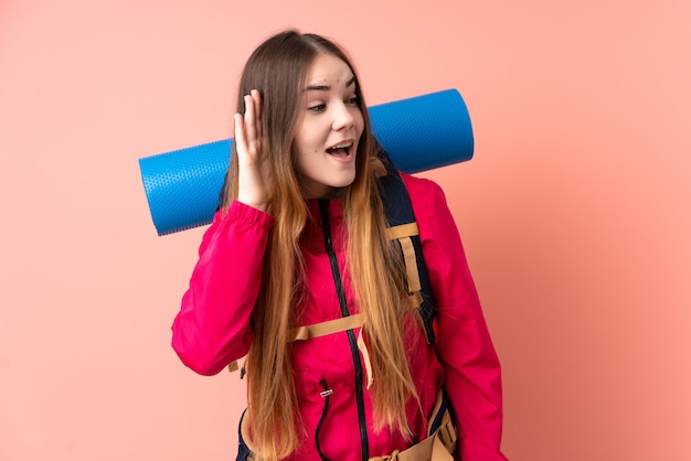 Junges Bergsteigermädchen mit einem großen Rucksack über rosa Wand, die etwas hört