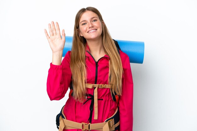 Junges Bergsteigermädchen mit großem Rucksack über isoliertem weißem Hintergrund, das mit der Hand mit fröhlichem Ausdruck salutiert