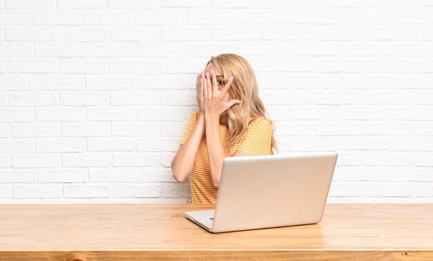 junges blondes Mädchen am Laptop, das Gesicht mit Händen bedeckt, die zwischen Fingern spähen