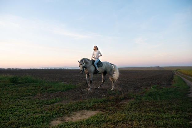 Junges blondes Mädchen, das auf einem Pferd auf dem Feld während des Sonnenuntergangs reitet
