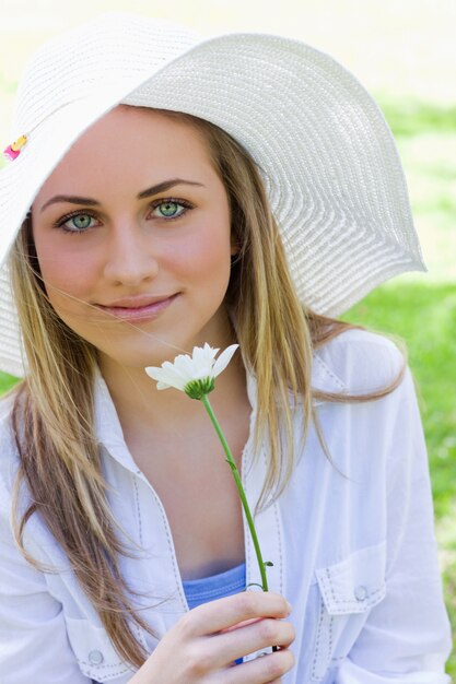 Junges blondes Mädchen, das einen weißen Hut beim Anhalten einer weißen Blume trägt