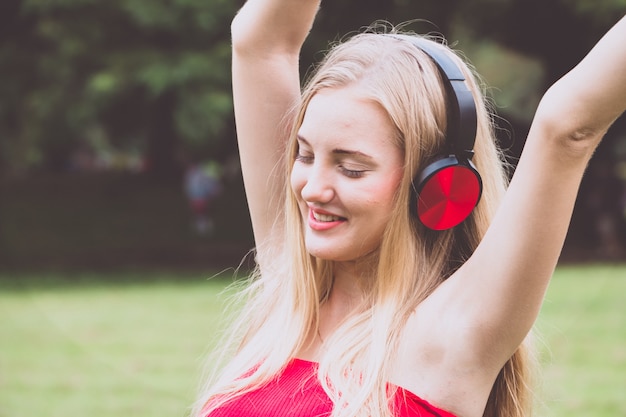 Junges blondes Mädchen, das glücklich mit Kopfhörern am Park lächelt.