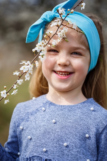 Junges blondes Mädchen, das in einem blühenden Garten steht. Blühende Kirsche. Porträt des schönen kleinen Mädchens. Nahaufnahme des kleinen Modellgesichts. Sakura blüht, Frühlingsabendzeit