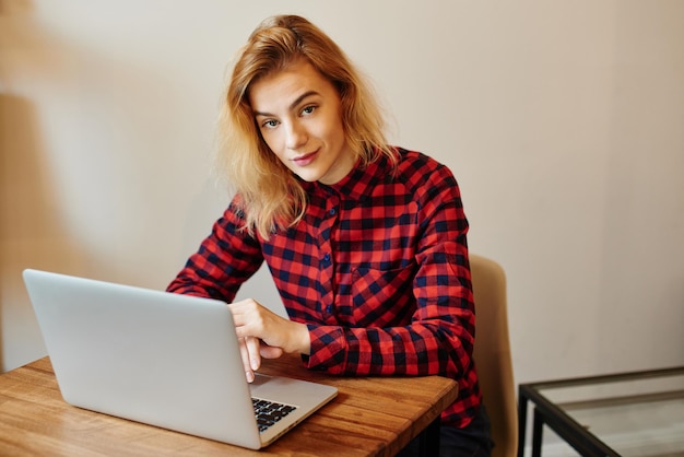 Junges blondes Mädchen, das mit einem Laptop in einem Cafétisch sitzt. Ein Student im roten Hemd läuft