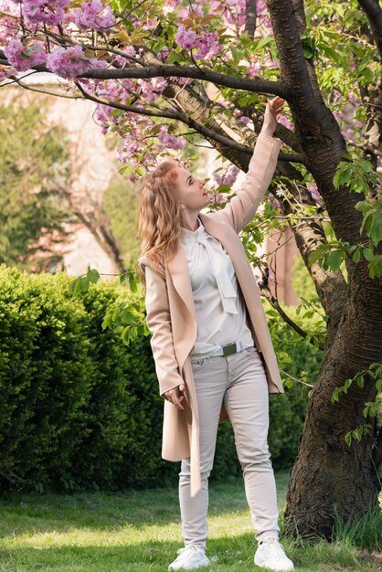 Junges blondes Mädchen geht im Frühlingspark in der Nähe des Kirschblütenbaums Sakura Vertikaler Rahmen