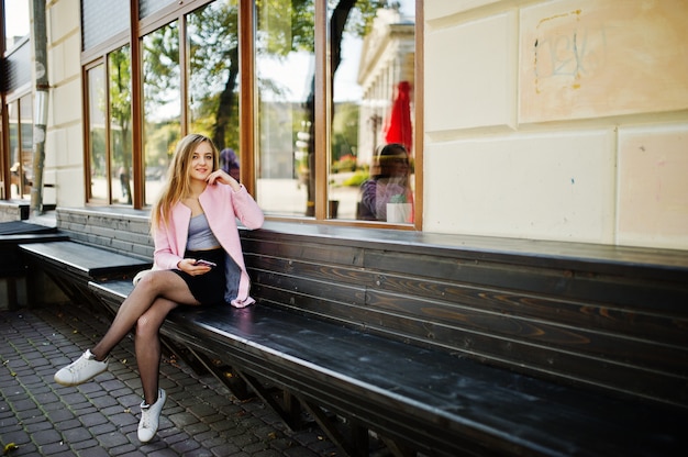 Junges blondes Mädchen im schwarzen Rock und im rosa Mantel, die auf Bank an der Stadt mit Handy an den Händen sitzt.