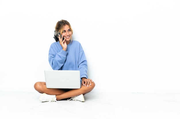 Junges blondes uruguayisches Mädchen mit dem Laptop lokalisiert auf weißer Wand, die ein Gespräch mit dem Mobiltelefon mit jemandem hält