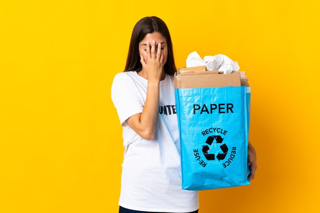 Junges brasilianisches Mädchen, das einen Recyclingbeutel voll Papier hält, um lokalisiert auf gelber Wand mit müde und krankem Ausdruck zu recyceln