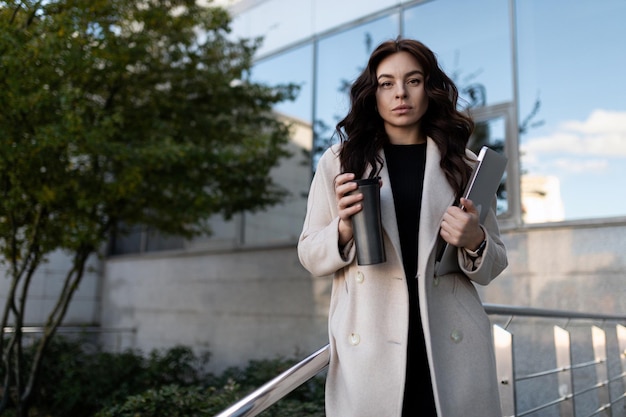 Junges brünettes europäisches Frauenmodell mit einem Tablet und einem Thermobecher auf dem Hintergrund eines Unternehmens