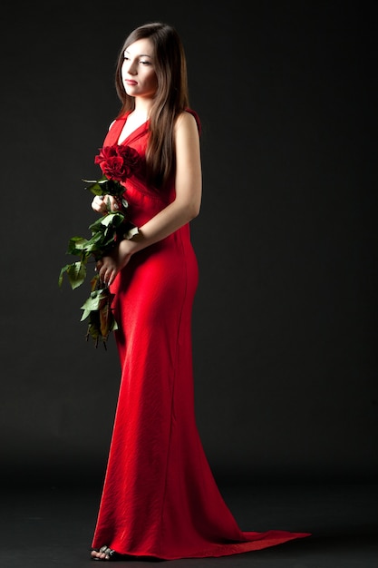 Junges brünettes Frauenmodell im roten langen Abendkleid stehend und hält Strauß der roten Rosen in den Händen über dunklem Hintergrund im Fotostudio