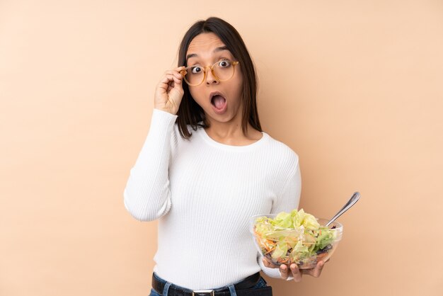 Junges brünettes Mädchen, das einen Salat über isolierter Wand mit Gläsern hält und überrascht