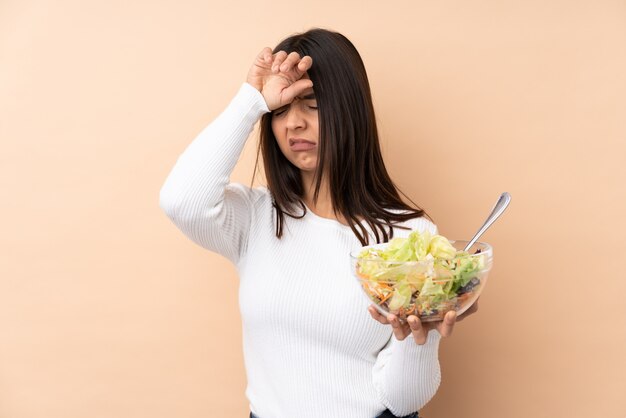 Junges brünettes Mädchen, das einen Salat über isolierter Wand mit müde und krankem Ausdruck hält