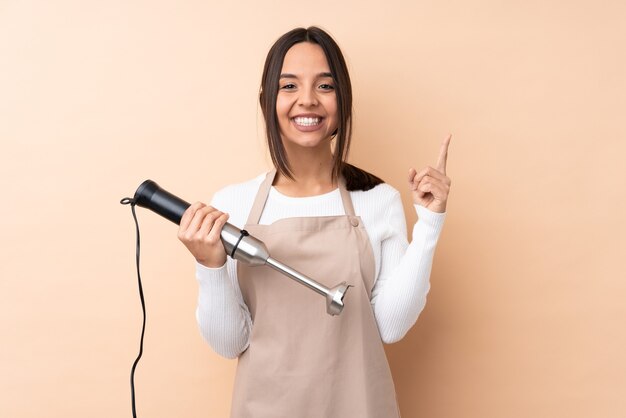 Junges brünettes Mädchen, das Handmixer über isoliertem Hintergrund verwendet, der eine große Idee aufzeigt