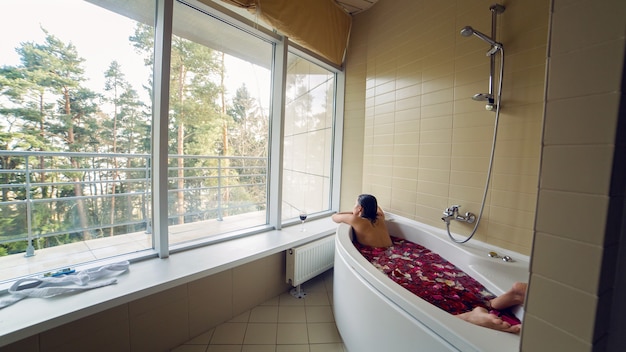 Junges brünettes mädchen mit langen haaren ist ein weißes bad mit rosenblättern und schaut aus dem großen fenster, hinter dem bäume stehen