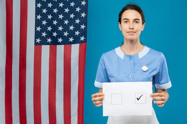 Junges brünettes Zimmermädchen in blauer Uniform zeigt Ihnen ihren Stimmzettel mit Zecke in einem der Quadrate, während Sie gegen US-Flagge stehen