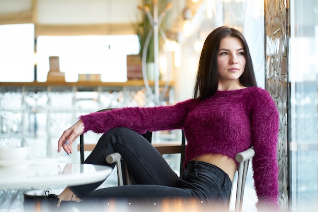 Junges Brunettemädchen, das auf Stuhl im Café sitzt und Fenster betrachtet