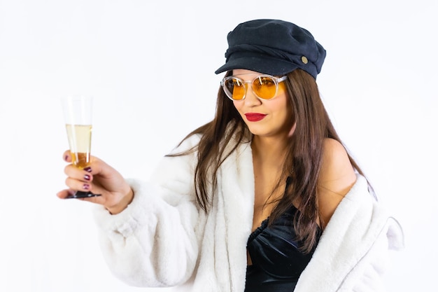 Junges Brunettemädchen im schwarzen Hut, das Silvester auf einem weißen Hintergrund feiert. Ich fordere das Model mit einem Glas Champagner heraus