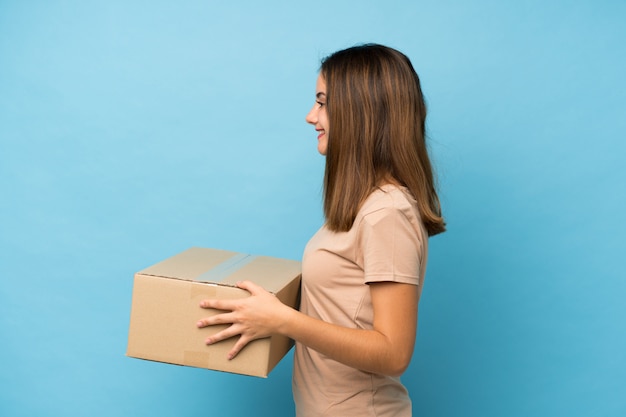 Junges Brunettemädchen über dem lokalisierten Blau, das einen Kasten hält, um ihn auf einen anderen Standort zu bewegen