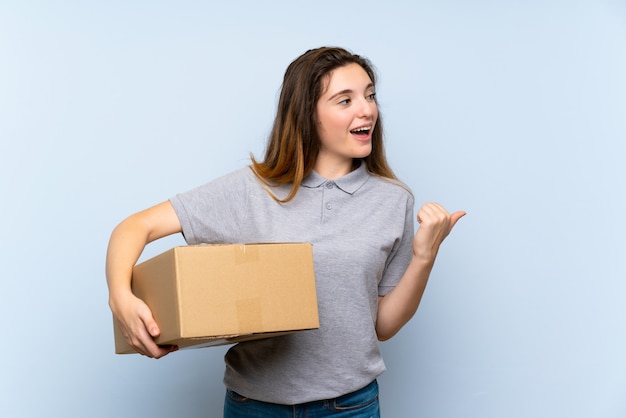 Junges Brunettemädchen über der lokalisierten blauen Wand, die einen Kasten hält, um ihn auf einen anderen Standort zu bewegen und Seite zu zeigen