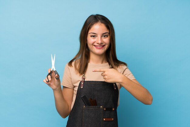 Junges Brunettemädchen über lokalisiertem Blau mit Friseur- oder Friseurkleid und Zeigen es