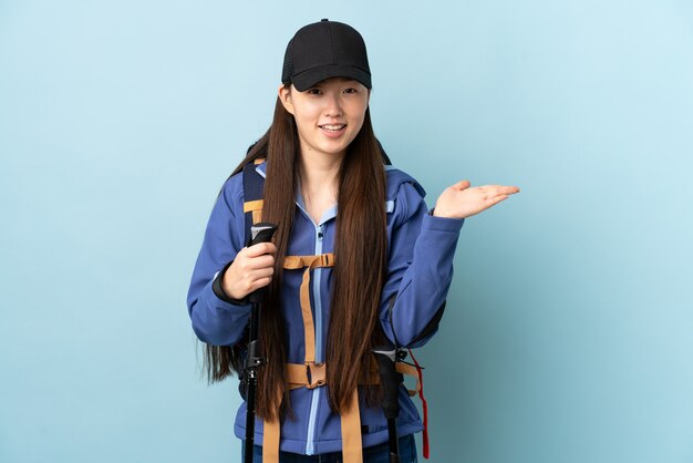 Junges chinesisches Mädchen mit Rucksack und Wanderstöcken über isolierter blauer Wand, die imaginären Copyspace auf der Handfläche hält, um eine Anzeige einzufügen