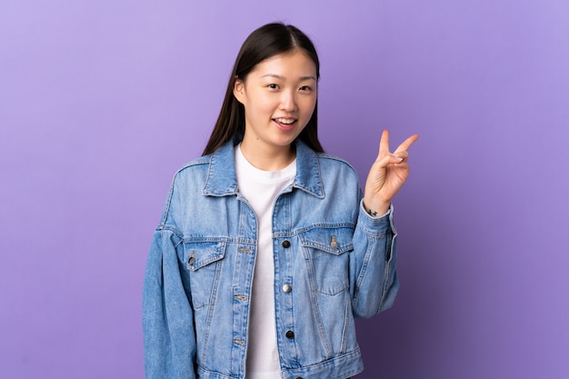 Junges chinesisches Mädchen über isolierte lila Wand lächelnd und Siegeszeichen zeigend