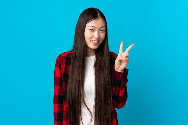 Junges chinesisches Mädchen über lokalisiertem blauem Hintergrund lächelnd und Siegeszeichen zeigend