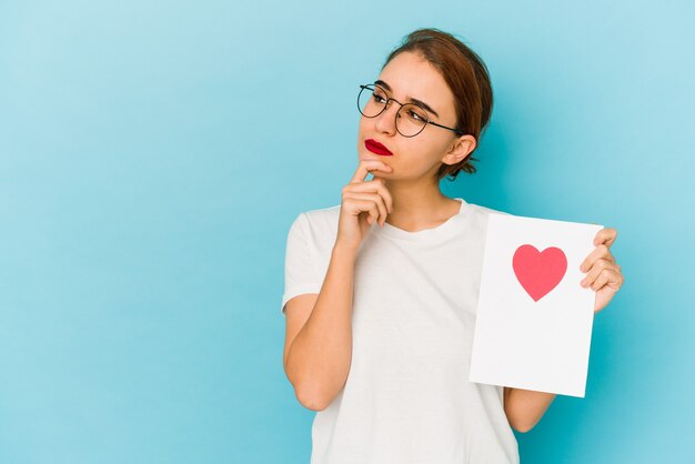 Junges dünnes arabisches Mädchen, das eine Valentinstagkarte hält, die seitwärts mit zweifelhaftem und skeptischem Ausdruck schaut.