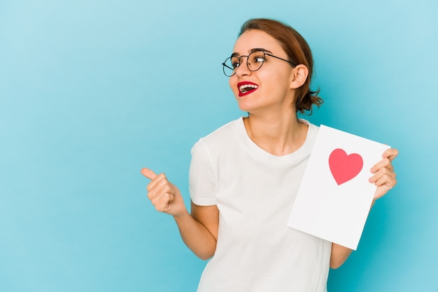 Junges dünnes arabisches Mädchen, das eine Valentinstagskarte hält, zeigt mit Daumenfinger weg, lachend und sorglos.