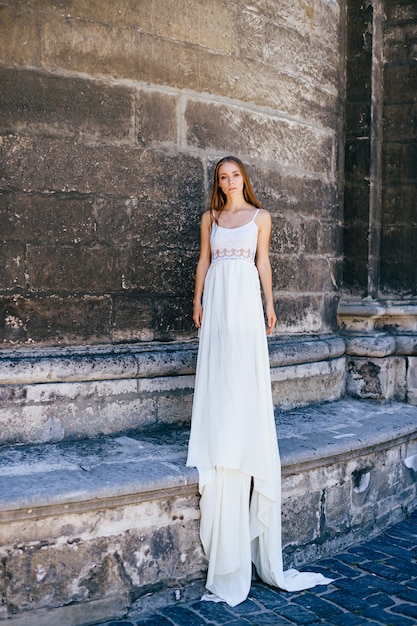 Junges elegantes Mädchen im langen weißen Kleid, das über alte Steinmauer aufwirft