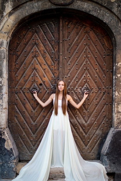 Junges elegantes Mädchen im langen weißen Kleid, das über der alten Tür aufwirft