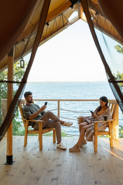 Junges erholsames Paar, das in Smartphones von Glamping House scrollt