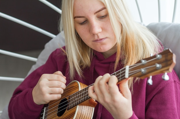 Junges erwachsenes blondes Mädchen, das lernt, Ukulele zu spielen, Online-Gitarrenlernen, selektiver Fokus